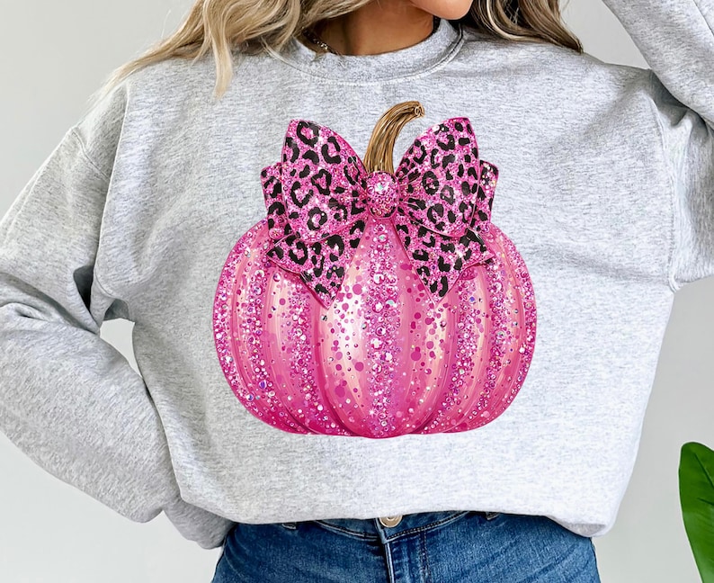 May include: A light gray sweatshirt featuring a large, pink glitter pumpkin with a leopard print bow. The pumpkin has a stem and is covered in pink glitter. The bow is black and brown.