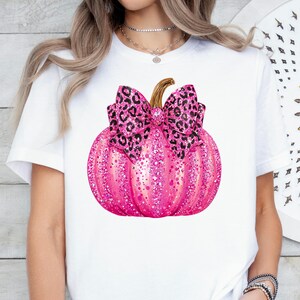 May include: A white t-shirt featuring a pink pumpkin design. The pumpkin is covered in glitter and has a leopard print bow. The stem is gold. A festive design for the fall season.