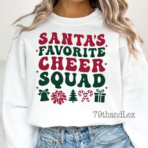 May include: White sweatshirt with the text "SANTA'S FAVORITE CHEER SQUAD" in red and green. The design includes festive icons like a bell, pom-poms, a Christmas tree, candy canes, and a gift box. The sweatshirt is a casual, holiday-themed apparel item.