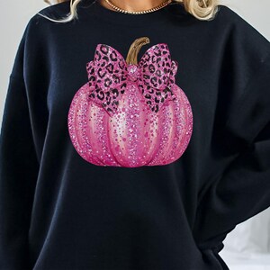 May include: Black sweatshirt featuring a pink glittery pumpkin with a leopard print bow. The pumpkin has a gold stem and is decorated with pink glitter. The sweatshirt is a solid black color.