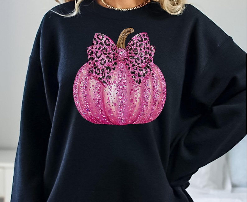 May include: Black sweatshirt featuring a pink glittery pumpkin with a leopard print bow. The pumpkin has a gold stem and is decorated with pink glitter. The sweatshirt is a solid black color.