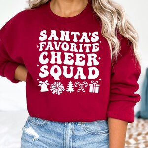 May include: A burgundy sweatshirt with the text "SANTA'S FAVORITE CHEER SQUAD" in white, along with festive icons. The design includes a bell, pom-pom, Christmas tree, candy cane, and a gift box. A cozy, seasonal top.
