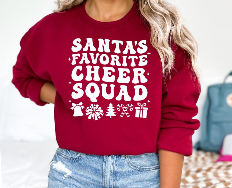 May include: A burgundy sweatshirt with the text "SANTA'S FAVORITE CHEER SQUAD" in white, along with festive icons. The design includes a bell, pom-pom, Christmas tree, candy cane, and a gift box. A cozy, seasonal top.