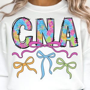 May include: White sweatshirt with the letters "CNA" in a tie-dye floral pattern. The letters are outlined in black with decorative stitching. Below the letters are three bows in pink, blue, and green.