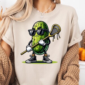 May include: A white t-shirt with a cartoon pickle wearing sunglasses and holding a lacrosse stick. The pickle is green with a yellow background.