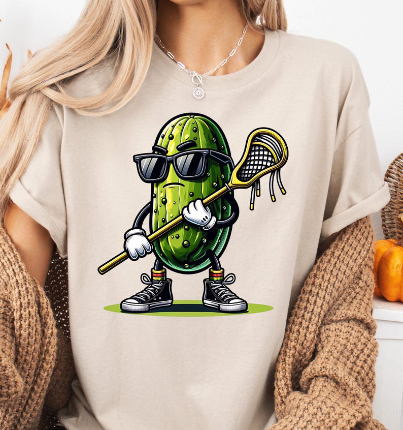 May include: A white t-shirt with a cartoon pickle wearing sunglasses and holding a lacrosse stick. The pickle is green with a yellow background.