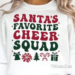 May include: White sweatshirt with the text "SANTA'S FAVORITE CHEER SQUAD" in red and green. The design includes festive icons like a bell, pom-pom, Christmas tree, candy canes, and a gift box. The brand name "79thandLex" is at the bottom.