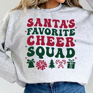May include: A light gray sweatshirt with the text "SANTA'S FAVORITE CHEER SQUAD" in red and green. The design includes a bell, pom-pom, Christmas tree, candy canes, and a gift box.