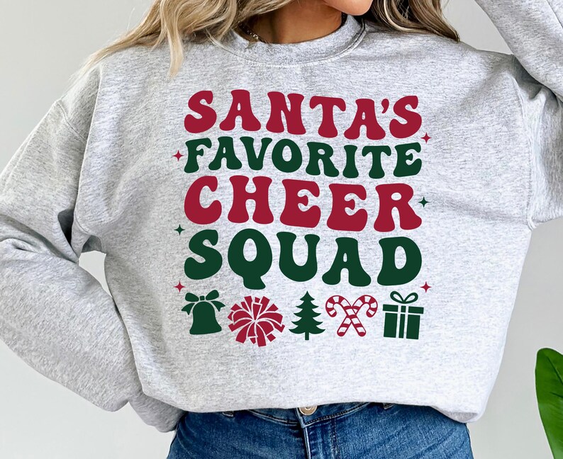 May include: A light gray sweatshirt with the text "SANTA'S FAVORITE CHEER SQUAD" in red and green. The design includes a bell, pom-pom, Christmas tree, candy canes, and a gift box.