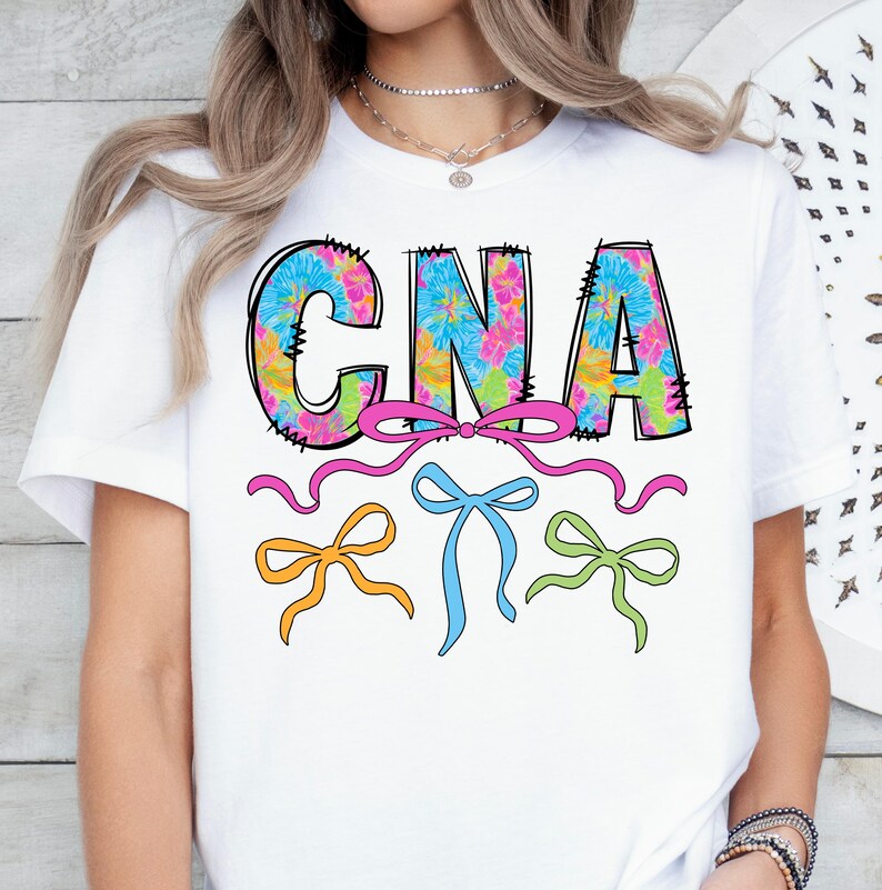 May include: White t-shirt with the letters "CNA" in a black outline, filled with a floral pattern in blue, pink, yellow, and green. Pink ribbon and three colorful bows in orange, blue, and green are below the letters.