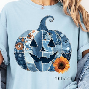 May include: A light blue t-shirt featuring a large pumpkin graphic composed of various blue and white floral and denim-style patchwork patterns. The pumpkin has a dark blue jack-o'-lantern face and a bright yellow sunflower at the base. Text reads 79thandLex.