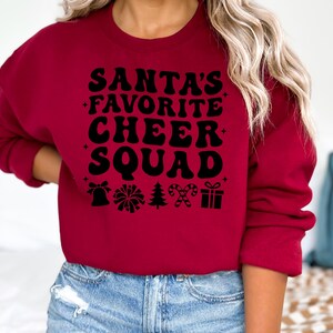 May include: A burgundy sweatshirt with the text "SANTA'S FAVORITE CHEER SQUAD" in black, along with festive icons. The design includes a bell, pom-pom, Christmas tree, candy canes, and a gift box. The sweatshirt is a crew neck style.