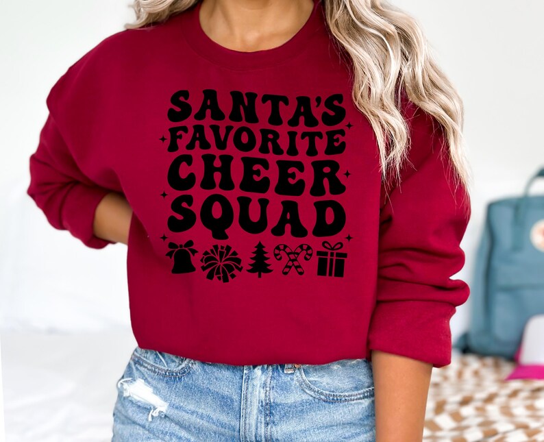 May include: A burgundy sweatshirt with the text "SANTA'S FAVORITE CHEER SQUAD" in black, along with festive icons. The design includes a bell, pom-pom, Christmas tree, candy canes, and a gift box. The sweatshirt is a crew neck style.