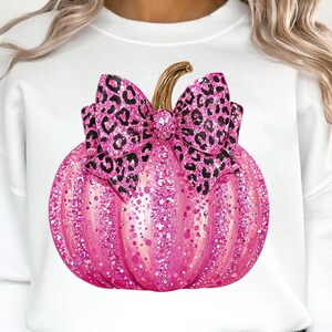 May include: A white sweatshirt featuring a pink glitter pumpkin with a leopard print bow. The pumpkin has a gold stem and is covered in sparkling glitter. The bow is a contrasting black and pink leopard print.