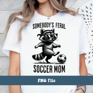 May include: White t-shirt with a black and white illustration of a raccoon wearing sunglasses and kicking a soccer ball. The text "Somebody's Feral Soccer Mom" is printed on the shirt.