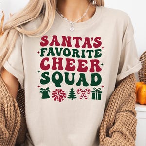 May include: Beige t-shirt with the text "SANTA'S FAVORITE CHEER SQUAD" in red and green. The design includes festive icons like a bell, pom-pom, Christmas tree, candy cane, and a gift box. A perfect holiday tee.