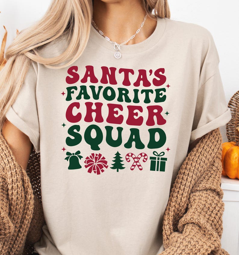 May include: Beige t-shirt with the text "SANTA'S FAVORITE CHEER SQUAD" in red and green. The design includes festive icons like a bell, pom-pom, Christmas tree, candy cane, and a gift box. A perfect holiday tee.