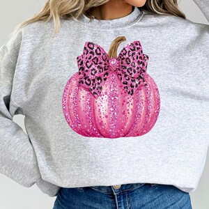 May include: A light gray sweatshirt featuring a large, pink glitter pumpkin with a leopard print bow. The pumpkin has a brown stem and is the focal point of the design. The sweatshirt is a casual, comfortable style.