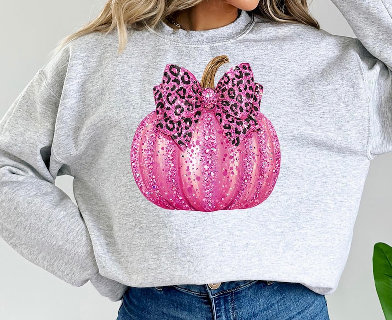 May include: A light gray sweatshirt featuring a large, pink glitter pumpkin with a leopard print bow. The pumpkin has a brown stem and is the focal point of the design. The sweatshirt is a casual, comfortable style.