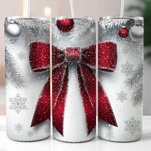 May include: A set of three stainless steel tumblers with a festive winter design. Each tumbler features a large red bow with a snowy effect, silver ornaments, snowflakes, and pine branches against a white background. The tumblers have clear straws.