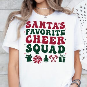 May include: White t-shirt with the text "SANTA'S FAVORITE CHEER SQUAD" in red and green. The design includes a bell, pom-pom, Christmas tree, candy cane, and gift box. A festive holiday-themed tee.