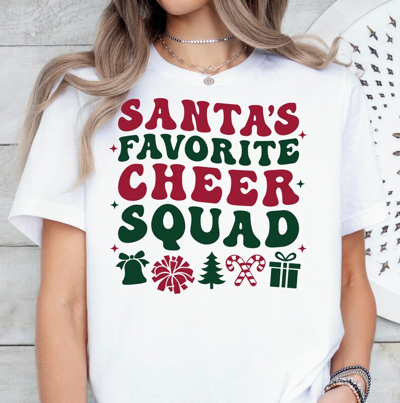 May include: White t-shirt with the text "SANTA'S FAVORITE CHEER SQUAD" in red and green. The design includes a bell, pom-pom, Christmas tree, candy cane, and gift box. A festive holiday-themed tee.