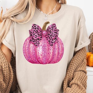 May include: A beige t-shirt featuring a pink glitter pumpkin with a leopard print bow. The pumpkin has a brown stem and is the focal point of the design. The shirt is a casual style.