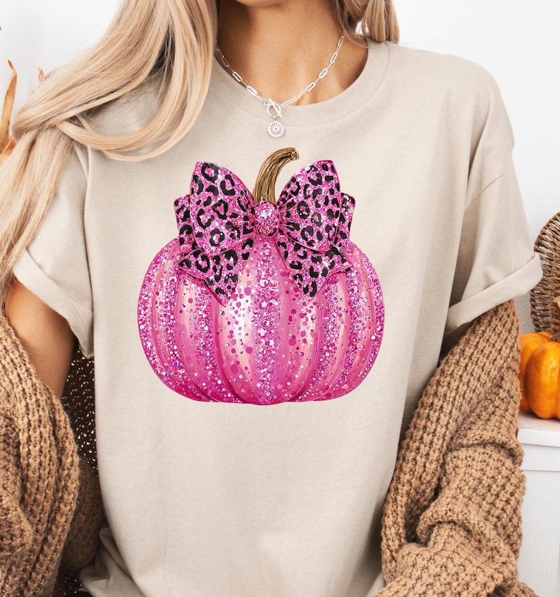 May include: A beige t-shirt featuring a pink glitter pumpkin with a leopard print bow. The pumpkin has a brown stem and is the focal point of the design. The shirt is a casual style.