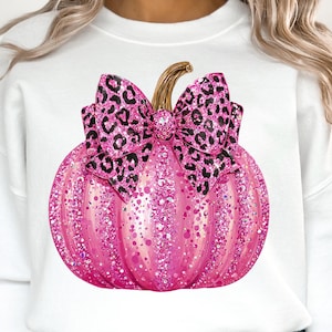 May include: A white sweatshirt featuring a pink glitter pumpkin with a leopard print bow. The pumpkin has a gold stem and is covered in sparkling glitter. The bow is a contrasting black and pink leopard print.