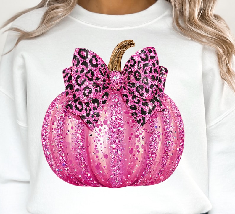 May include: A white sweatshirt featuring a pink glitter pumpkin with a leopard print bow. The pumpkin has a gold stem and is covered in sparkling glitter. The bow is a contrasting black and pink leopard print.