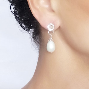 May include: A close-up of a silver earring with a clear, round gemstone at the top and a teardrop-shaped white pearl pendant. The earring is dangling from the ear of a person.