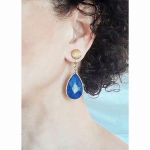 Könnte beinhalten: Goldene Tropfenohrringe mit einem blauen Edelstein. Die Ohrringe haben oben eine strukturierte goldene Kugel.