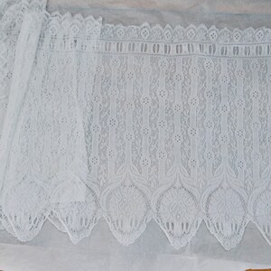 May include: White lace trim with a scalloped edge and a delicate floral pattern. The lace has a repeating pattern of vertical lines with small circles and a larger floral design at the bottom.