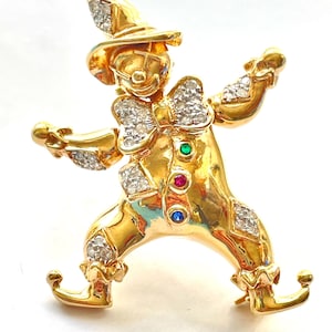 May include: A gold-toned clown brooch featuring a polished finish, adorned with clear sparkling rhinestones on the hat, bow tie, cuffs, and legs. The torso is decorated with three small round inset stones in green, red, and blue.
