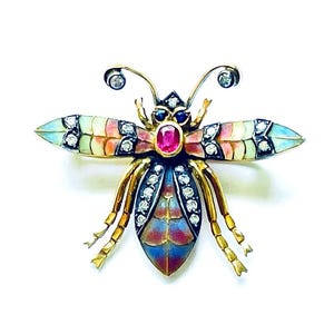 May include: A gold-toned insect brooch with a ruby center and diamond accents. The wings are multi-colored with blue, pink, and yellow hues. The brooch has a detailed design and is set against a white background.