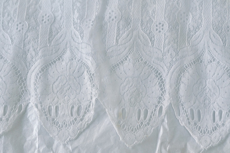 May include: White lace fabric with a delicate floral pattern. The lace has a scalloped edge.