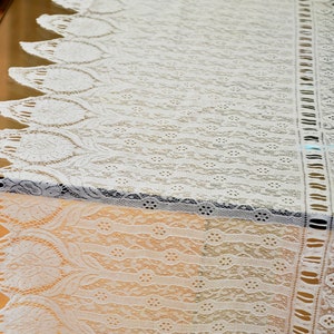 May include: White lace table runner with a delicate floral pattern. The runner has a scalloped edge and a fringe along the sides.