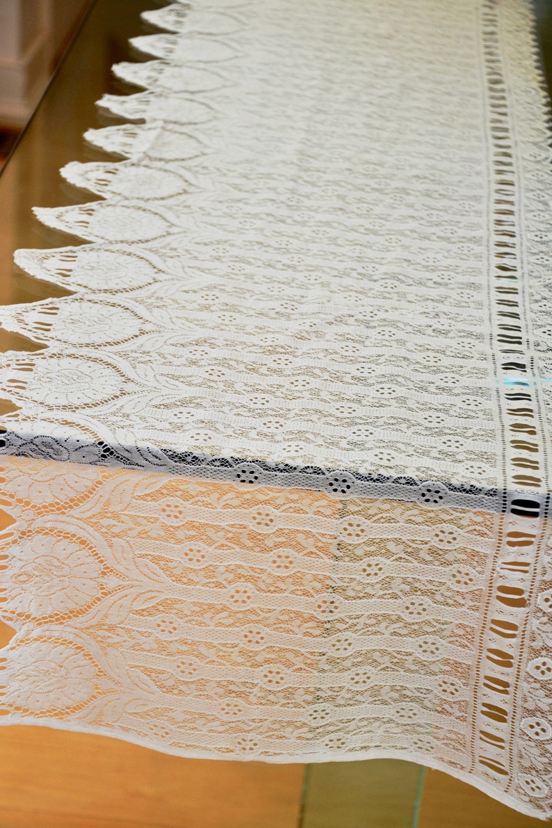 May include: White lace table runner with a delicate floral pattern. The runner has a scalloped edge and a fringe along the sides.