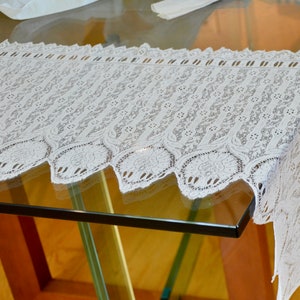 May include: White lace table runner with a scalloped edge. The runner is made of delicate lace with a floral pattern.