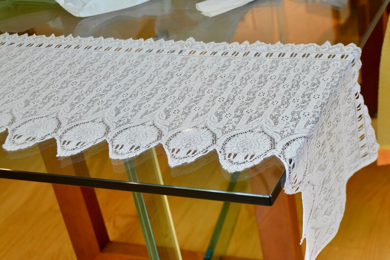 May include: White lace table runner with a scalloped edge. The runner is made of delicate lace with a floral pattern.