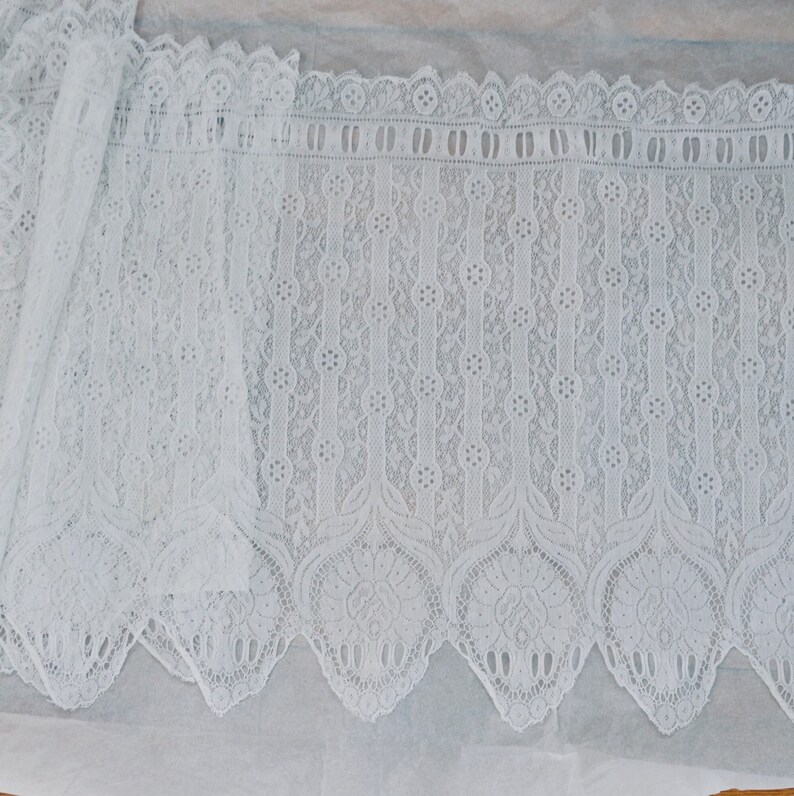 May include: White lace trim with a scalloped edge and a delicate floral pattern. The lace has a repeating pattern of vertical lines with small circles and a larger floral design at the bottom.