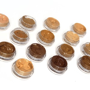 May include: An assortment of small, clear jars filled with various shades of foundation makeup. The colors range from light beige to deep brown, showcasing a diverse palette for different skin tones. The jars are arranged on a white surface.