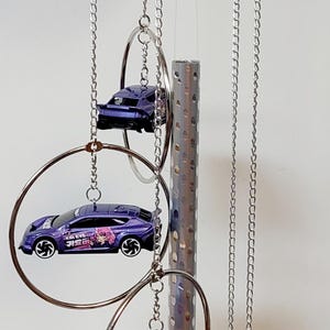 May include: A silver wind chime with four purple toy cars hanging from it. The cars have a cartoon girl and the words "K/DA EVS" printed on them.