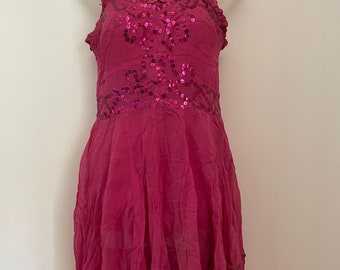 May include: A magenta-colored dress with a flowing skirt and spaghetti straps. The bodice is adorned with sequins, and the straps have a ruffled trim. The dress is displayed on a white mannequin, showcasing its design and style.