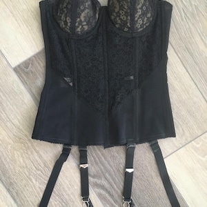 May include: Black vintage corset with lace detailing on the cups and front panel. The corset has adjustable straps and garter belt attachments. The garment is made of a smooth, matte fabric.