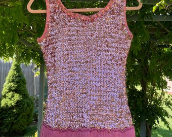 May include: A sleeveless, pink sequined dress with a fringe hemline. The dress is hanging on a wooden hanger against a backdrop of green foliage and grass. The sequins shimmer, and the fringe is a darker shade of pink.
