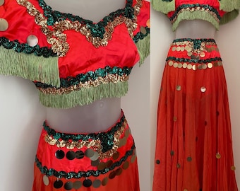 May include: A vibrant orange belly dance costume with a matching top and skirt. The top features green fringe, gold and green sequin trim, and gold coin accents. The skirt is adorned with gold coins and a green and gold sequin waistband.
