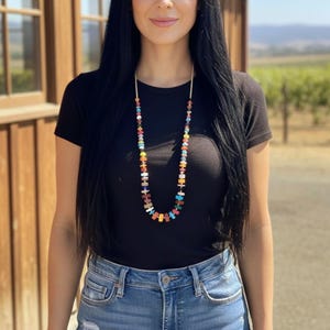 May include: A long, colorful beaded necklace with a gold chain. The necklace features a variety of rectangular and round beads in shades of red, blue, yellow, and green. The person is wearing a black t-shirt and blue jeans.