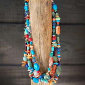 Op de afbeelding: Een meerrijige ketting gemaakt van verschillende gekleurde stenen, waaronder turquoise, koraal en lapis lazuli. De ketting wordt getoond op een houten standaard.