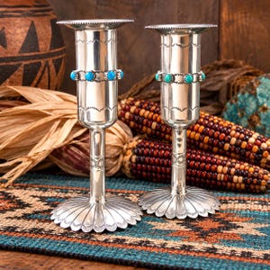 May include: Two silver candlesticks featuring a cylindrical body with a flared rim and a wide, petal-shaped base. Each candlestick is decorated with a band of small, round, bright blue turquoise stones and intricate stamped metal patterns.